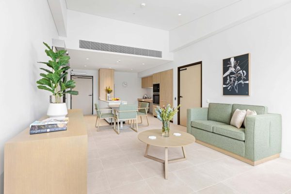 Lidcombe SDA 6 A modern, minimalistic living and dining area with light wood furniture, green sofa, plants, and a wall-mounted artwork in a bright, neutral-toned room.