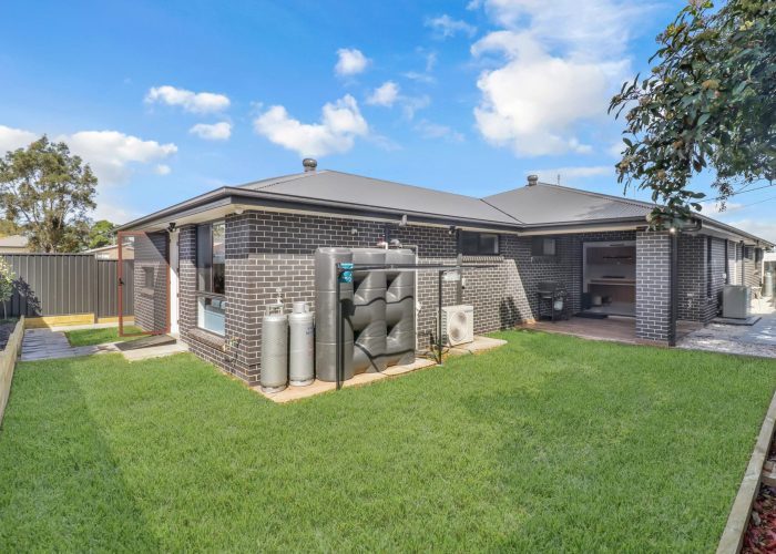 CHESTERHILL 2 4 Modern brick house exterior with a neat lawn, air conditioning unit, water tanks, and gas cylinders on the side. The sky is partly cloudy.