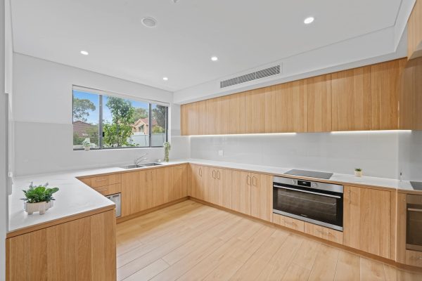 A modern kitchen with wooden cabinets, white countertops, stainless steel appliances, and a window overlooking trees. Light wood flooring complements the warm tones.