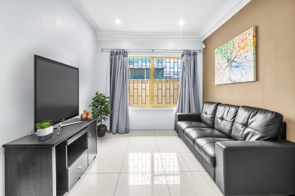 Fairfield 2 Modern living room with a black leather sofa, wall-mounted TV, potted plant, tiled floor, and a large window with gray curtains and bars. A colorful tree painting hangs on the wall.
