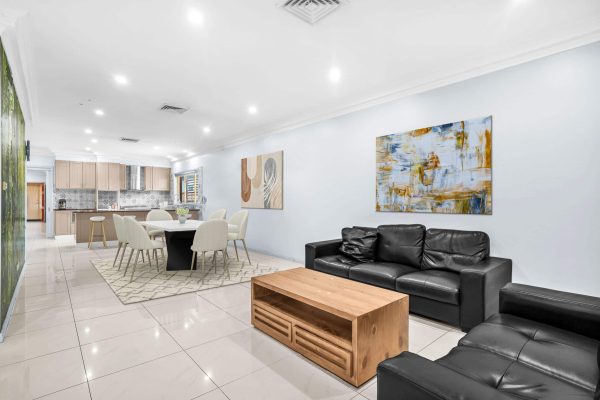 Fairfield 3 Modern open-plan living and dining area with black leather sofas, wooden coffee table, dining table with six chairs, kitchen in background, and abstract artwork on white walls.