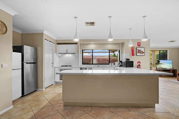 Kellyville 4 Modern kitchen with white countertops, stainless steel appliances, pendant lights, and a large window overlooking a garden. Adjacent living area with television is partially visible.