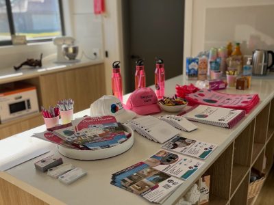 A kitchen island displays promotional items, brochures, pink hats, pens, water bottles, snacks, and drinks, with a microwave and kettle visible in the background.