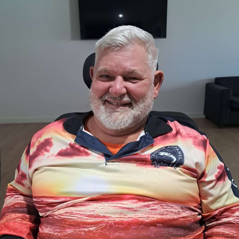 Success Story: Gary’s New Chapter at Walker Street, Merrylands 1 Older man with white hair and beard sitting indoors, smiling, wearing a colorful shirt with a seascape design. A black TV is mounted on the wall behind him and chairs are visible in the background.