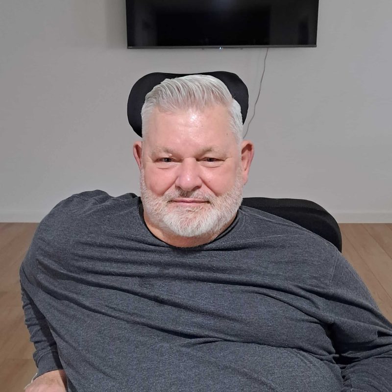 Success Story: Gary’s New Chapter at Walker Street, Merrylands 3 An older man with short white hair and a beard is sitting in a chair, wearing a grey long-sleeve shirt, with a TV mounted on the wall behind him.
