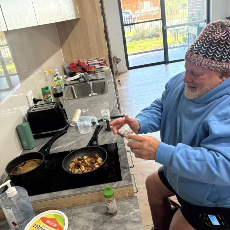 Success Story: Gary’s New Chapter at Walker Street, Merrylands 2 A man in a blue hoodie seasons food cooking in pans on a kitchen stove, with various ingredients and utensils on the counter.