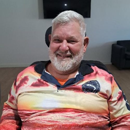 An older man with white hair and a beard is sitting indoors, smiling. He wears a colorful shirt with a landscape print and logo—an inspiring SDA participant story reflecting NDIS success in Beyond Care SDA homes.