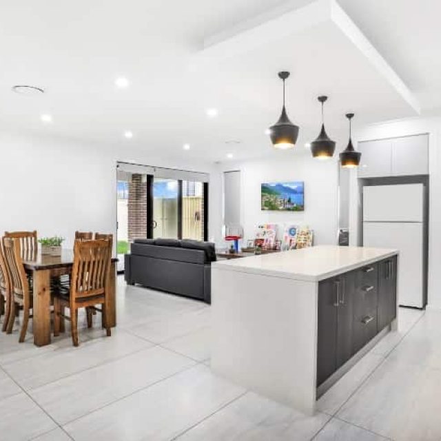 Modern open-plan kitchen and living area with white walls, island counter, hanging lights, dining table, chairs, and a couch facing a wall-mounted TV next to glass doors.