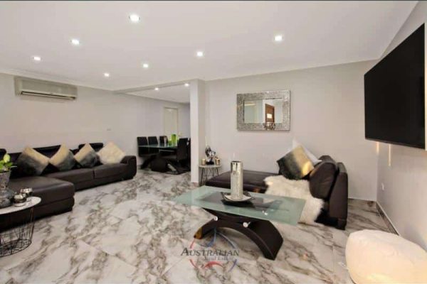 Modern living room with marble floors, two black leather sofas, a glass coffee table, and a large wall-mounted TV. A dining area is visible in the background.
