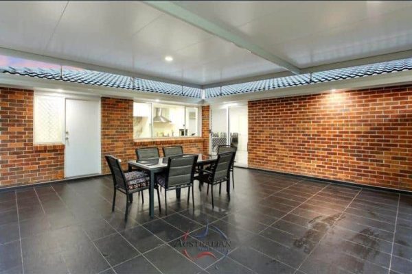 Outdoor patio with a table and four chairs on a tiled floor, surrounded by brick walls and covered by a roof. Connected to the indoors through windows and doors.