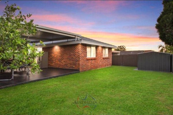 Brick house with a covered patio and lush green lawn, set against a colorful sunset sky. A tree and a shed are visible in the backyard.
