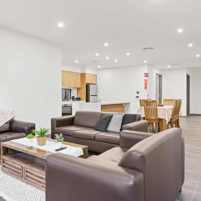 Modern open-plan living room and kitchen featuring brown leather sofas, a dining table, and neutral decor. Bright lighting and a minimalistic style throughout make the space welcoming for those seeking accessible living with NDIS services.