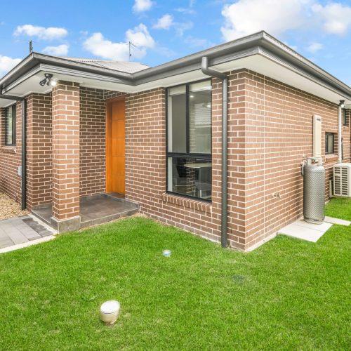 Single-story brick house with a brown door, paved walkway, green lawn, and outdoor air conditioning unit—ideal SIL Accommodation in Sydney with comfort and convenience under a cloudy blue sky.